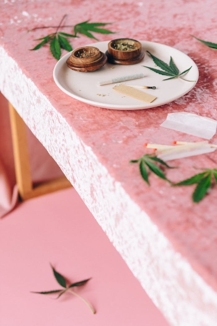 Cannabis leaves and rolling accessories on a pink textured surface, portraying a relaxed atmosphere.